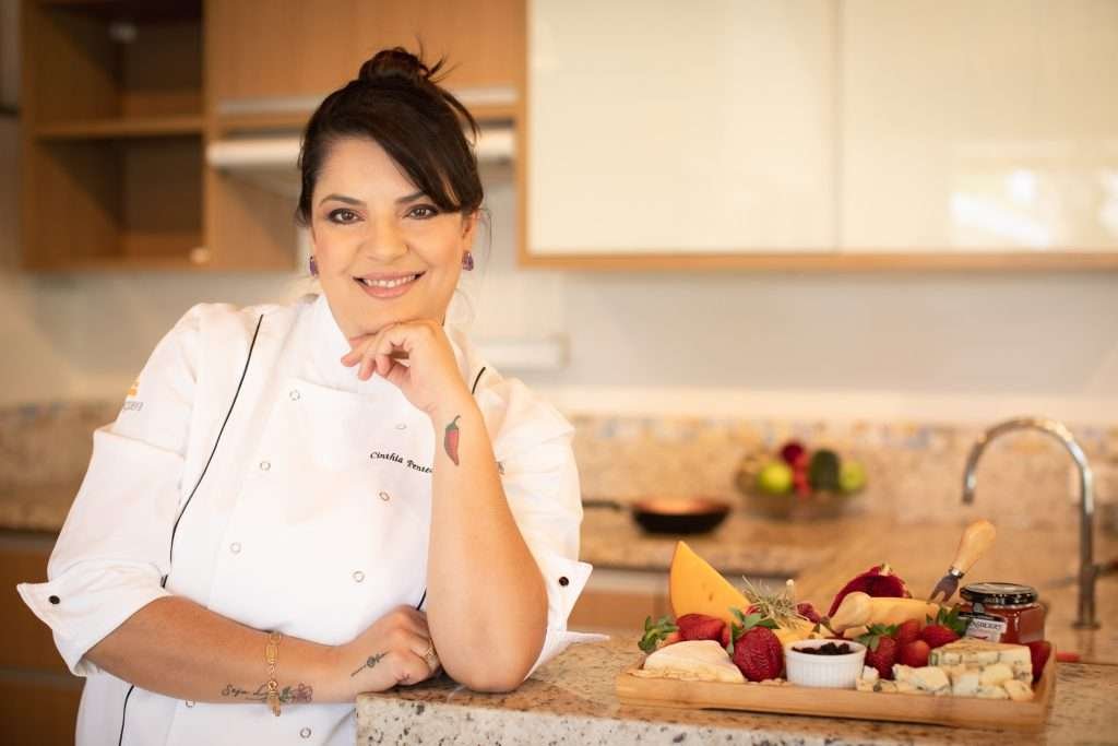 Cinthia apoiando o queixo na mão esquerda e o cotovelo esquerdo no balcão da cozinha ao lado de uma bandeja de frutas, queijo e geleia