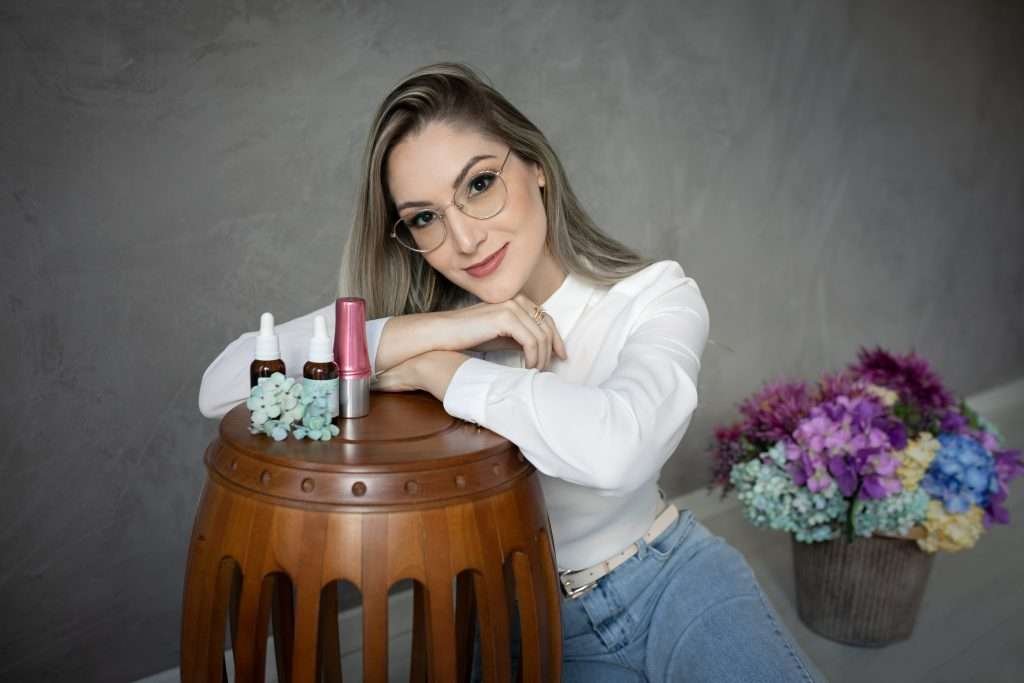 Diane de óculos sentada ao lado de um estande exibindo pequenas garrafas