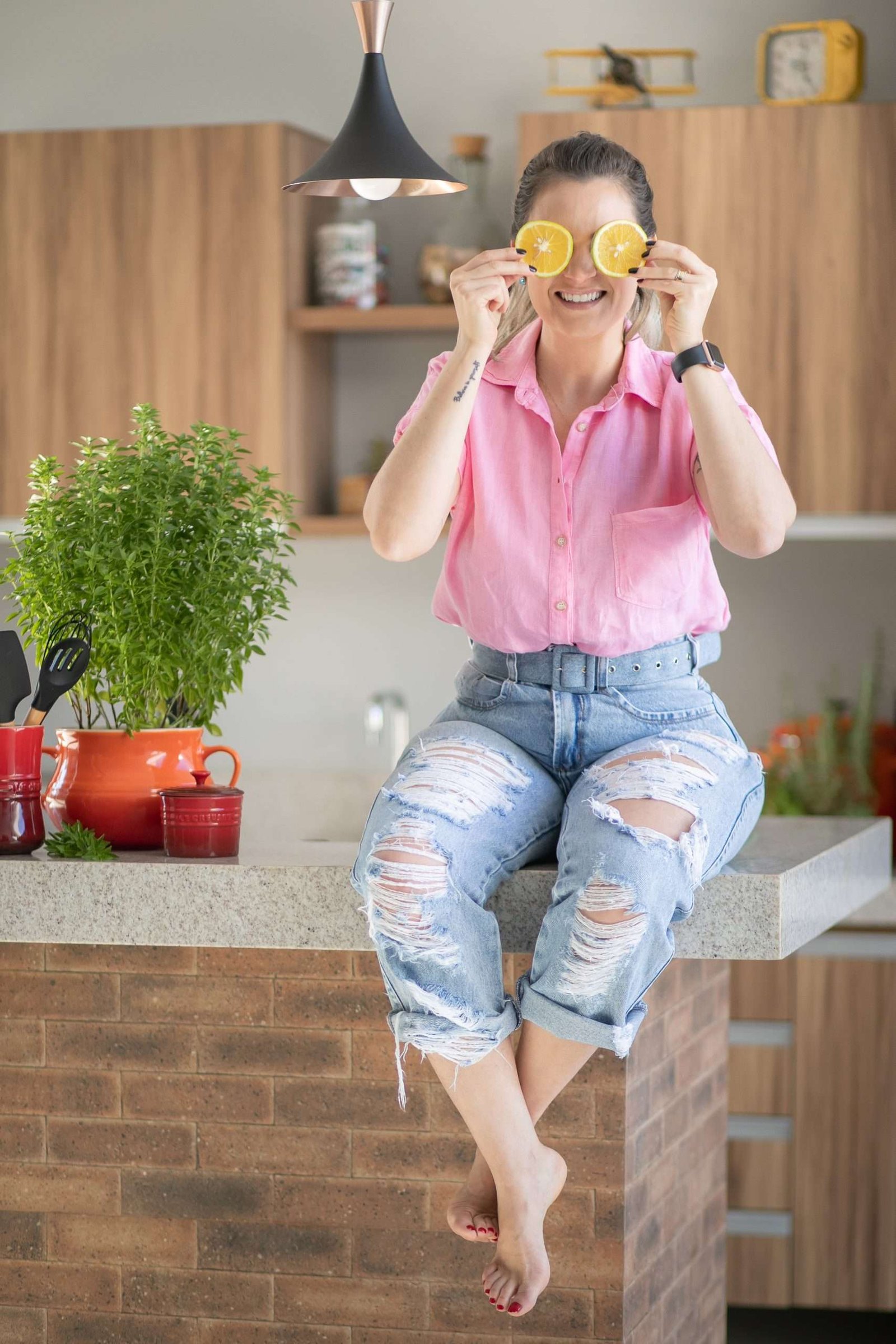 Nathassia segurando fatias de laranja sobre os olhos enquanto está sentada no balcão da cozinha