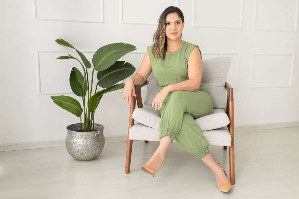 Paula sentada com as pernas cruzadas em uma cadeira com uma grande planta ao lado dela