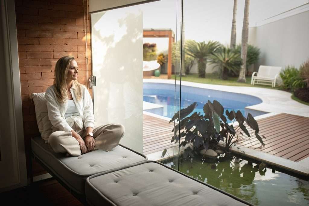 Renata pensando sentada em um sofá em frente a uma grande janela