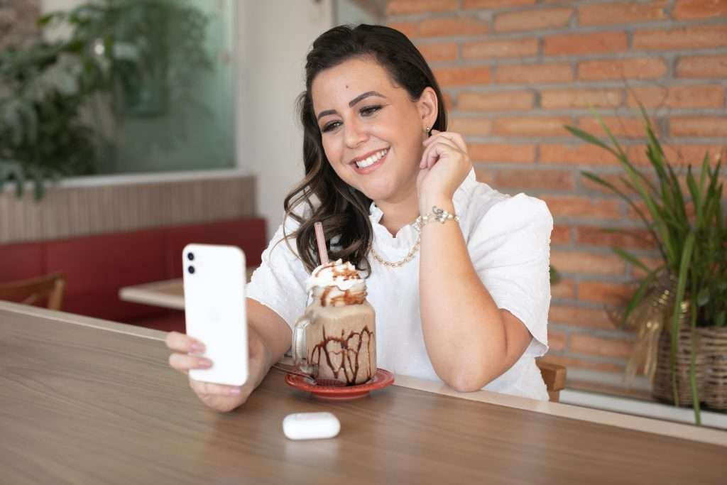 Ju sentada segurando seu celular tocando sua orelha com uma bebida de sobremesa na frente dela