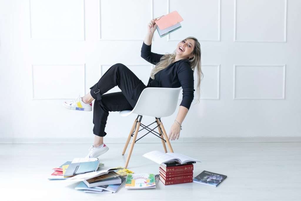 Mariana sorrindo alegremente sentada em uma cadeira branca com a cabeça para trás e a mão segurando um livro no ar