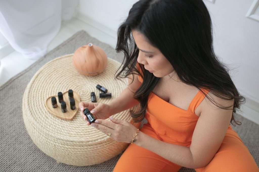 Mariana vestindo roupa laranja sentada no chão pernas cruzadas ao lado de pé com óleos segurando uma pequena garrafa nas mãos